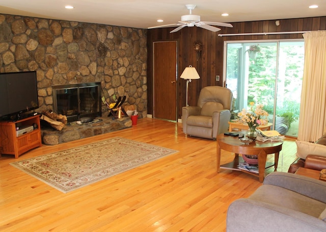 living room featuring ceiling fan, a stone fireplace, light wood-type flooring, and wooden walls