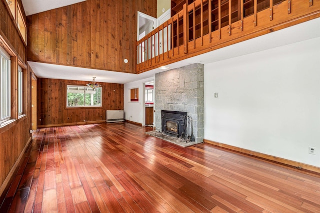 unfurnished living room with a wood stove, a baseboard radiator, a high ceiling, wood-type flooring, and wooden walls