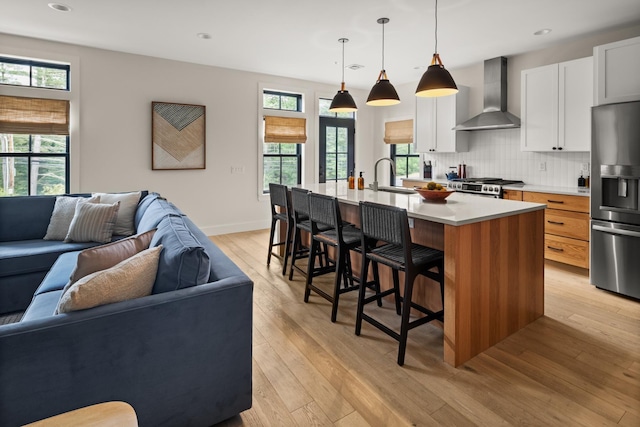 kitchen featuring white cabinetry, wall chimney range hood, decorative light fixtures, a center island with sink, and appliances with stainless steel finishes