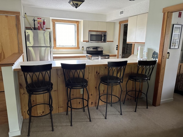 kitchen with stainless steel appliances, kitchen peninsula, cream cabinetry, a kitchen bar, and light carpet