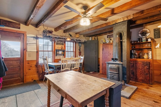 dining space with beam ceiling, a wood stove, ceiling fan, and wood walls