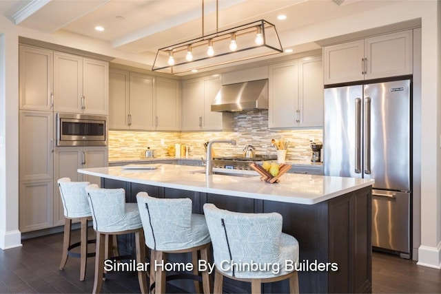 kitchen featuring tasteful backsplash, a center island with sink, built in appliances, and wall chimney range hood