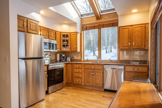 kitchen featuring sink, light hardwood / wood-style floors, decorative backsplash, vaulted ceiling with skylight, and appliances with stainless steel finishes