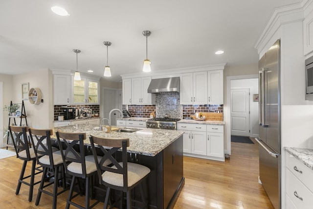 kitchen with white cabinetry, wall chimney exhaust hood, a center island with sink, appliances with stainless steel finishes, and light wood-type flooring