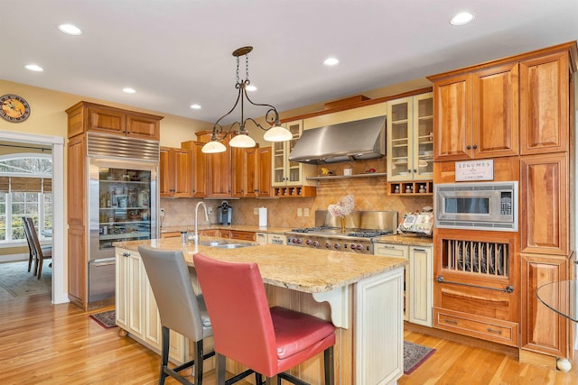 kitchen featuring hanging light fixtures, wall chimney exhaust hood, built in appliances, and an island with sink