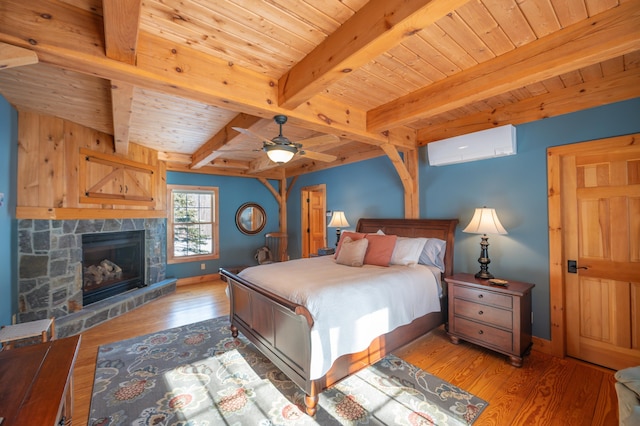 bedroom featuring ceiling fan, an AC wall unit, hardwood / wood-style floors, beam ceiling, and a stone fireplace