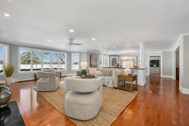 living room featuring ceiling fan with notable chandelier, a baseboard radiator, wood-type flooring, ornate columns, and crown molding