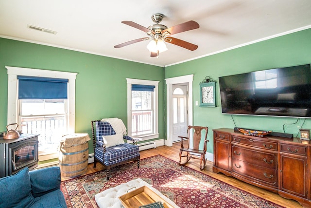 living room featuring ceiling fan, a wood stove, light wood-type flooring, baseboard heating, and ornamental molding