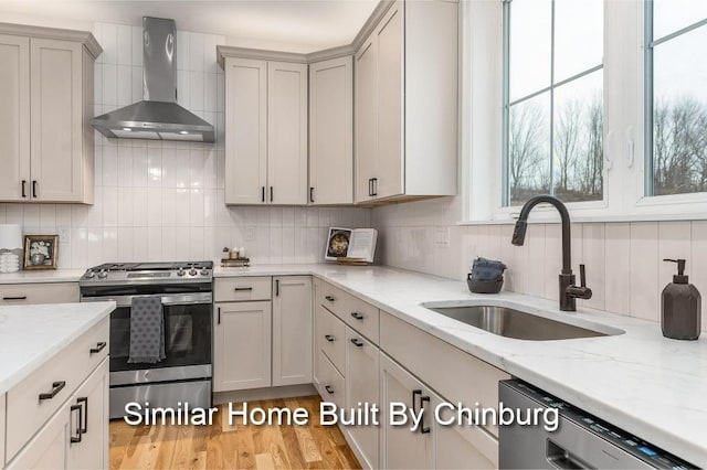 kitchen featuring wall chimney exhaust hood, sink, light stone counters, stainless steel appliances, and decorative backsplash