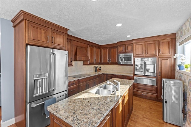 kitchen featuring appliances with stainless steel finishes, sink, light stone counters, wall chimney range hood, and a center island with sink