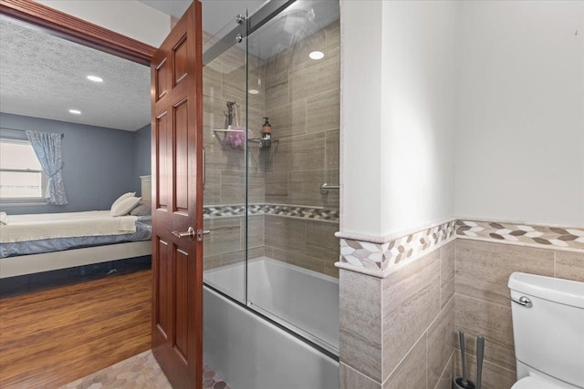 bathroom with a textured ceiling, bath / shower combo with glass door, toilet, and tile walls