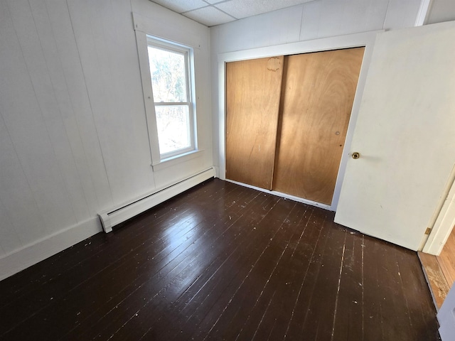 unfurnished bedroom with baseboard heating, a paneled ceiling, dark wood-type flooring, and a closet