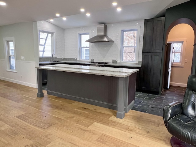 kitchen featuring light stone countertops, light wood-type flooring, a kitchen island, and wall chimney range hood