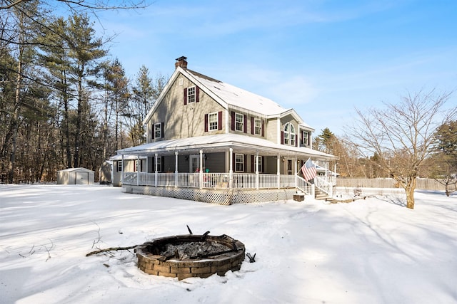 snow covered back of property featuring a porch, a storage unit, and an outdoor fire pit