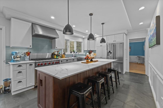 kitchen with wall chimney range hood, appliances with stainless steel finishes, white cabinetry, a kitchen island, and decorative light fixtures