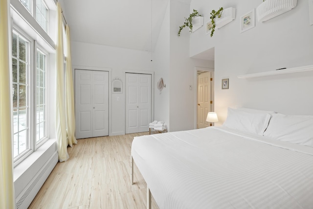 bedroom with high vaulted ceiling, light hardwood / wood-style floors, multiple closets, and baseboard heating