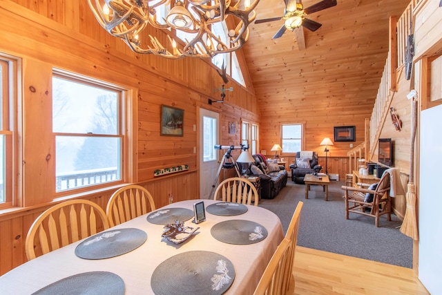 dining room featuring ceiling fan with notable chandelier, high vaulted ceiling, a fireplace, wooden walls, and light wood-type flooring