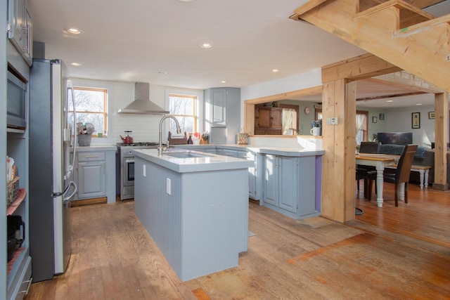 kitchen featuring sink, stainless steel appliances, light hardwood / wood-style floors, a center island with sink, and wall chimney exhaust hood