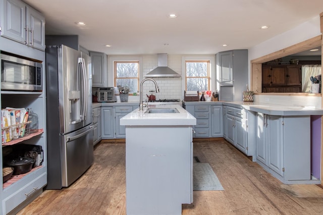 kitchen with light hardwood / wood-style flooring, a center island with sink, appliances with stainless steel finishes, decorative backsplash, and wall chimney range hood