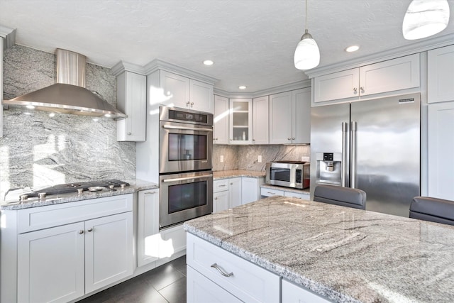 kitchen with appliances with stainless steel finishes, decorative light fixtures, white cabinets, light stone countertops, and wall chimney exhaust hood