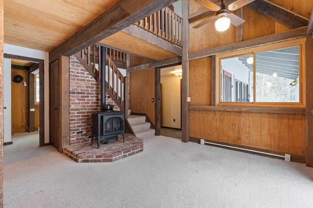 unfurnished living room featuring carpet flooring, wooden walls, a wood stove, and a baseboard heating unit