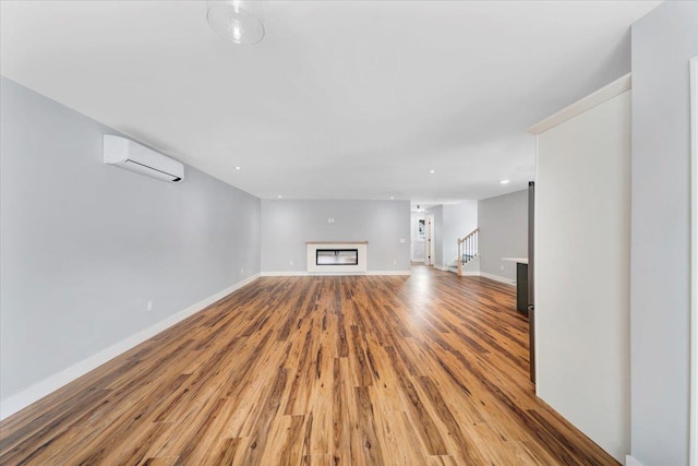 unfurnished living room with a wall mounted AC, a glass covered fireplace, light wood-style floors, baseboards, and stairs