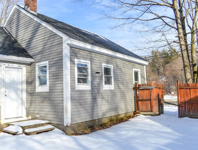 view of snow covered exterior featuring a shingled roof, fence, and a chimney