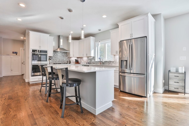 kitchen with hanging light fixtures, wall chimney range hood, a kitchen island, stainless steel appliances, and white cabinets