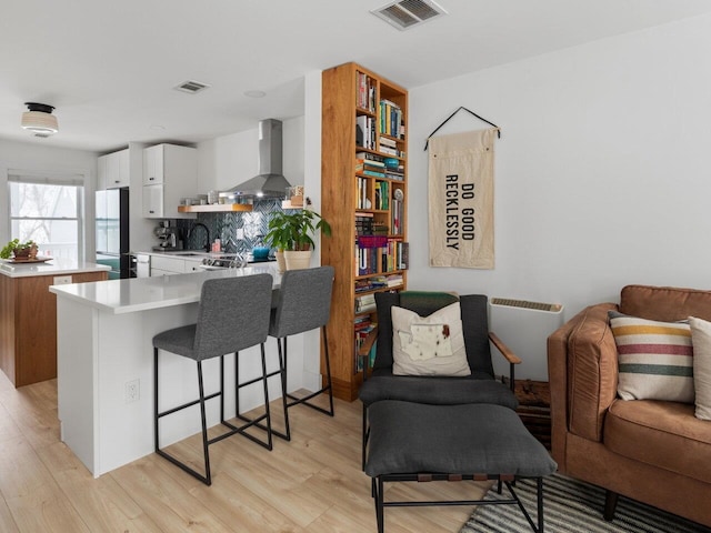 kitchen featuring wall chimney exhaust hood, fridge, kitchen peninsula, light hardwood / wood-style floors, and white cabinets