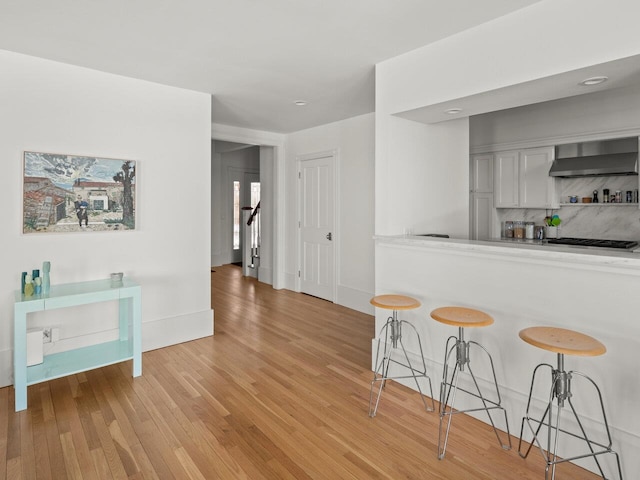 kitchen featuring a breakfast bar, backsplash, white cabinets, wall chimney range hood, and light wood-type flooring