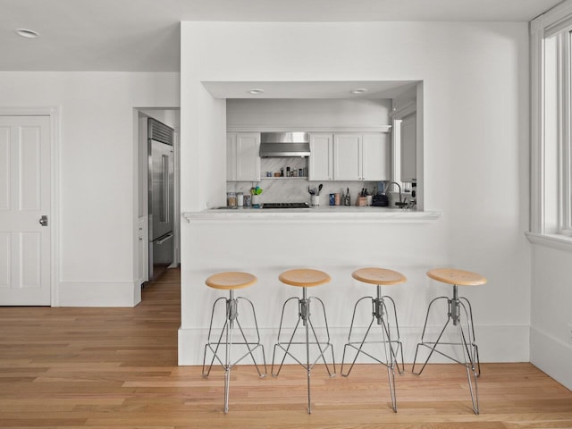 kitchen with built in fridge, white cabinetry, a kitchen breakfast bar, light wood-type flooring, and wall chimney exhaust hood