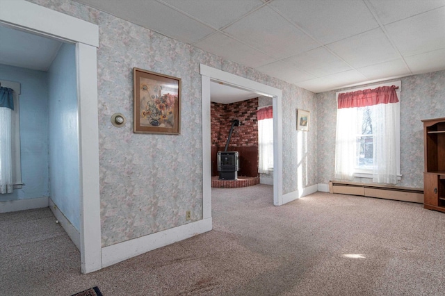 unfurnished living room featuring a drop ceiling, carpet floors, a wood stove, and a baseboard heating unit