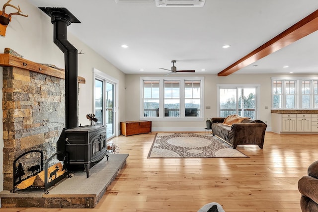 living room featuring ceiling fan, beam ceiling, light wood-type flooring, and a wood stove