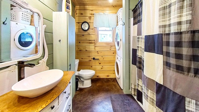 bathroom with stacked washer and dryer, wooden walls, vanity, concrete floors, and toilet