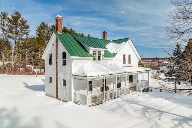 snow covered back of property with metal roof, covered porch, a chimney, and a standing seam roof