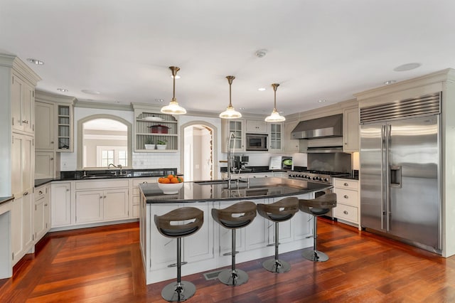 kitchen with pendant lighting, wall chimney range hood, dark hardwood / wood-style floors, built in appliances, and a center island with sink