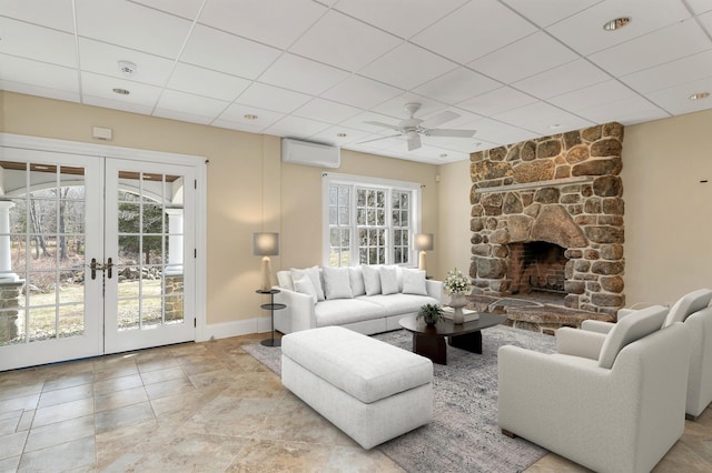 living room with french doors, a stone fireplace, a wall unit AC, ceiling fan, and a drop ceiling