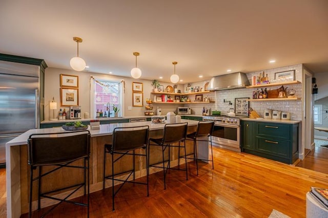 kitchen featuring green cabinets, hanging light fixtures, wall chimney range hood, a kitchen breakfast bar, and premium appliances