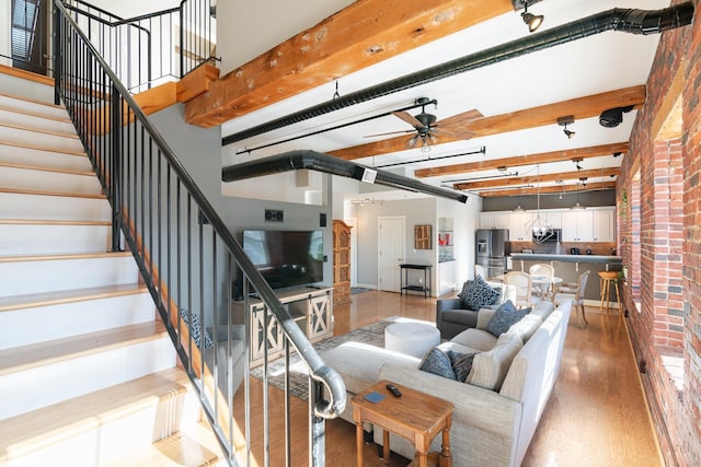 living room with light wood-style flooring, a towering ceiling, ceiling fan, beamed ceiling, and stairs