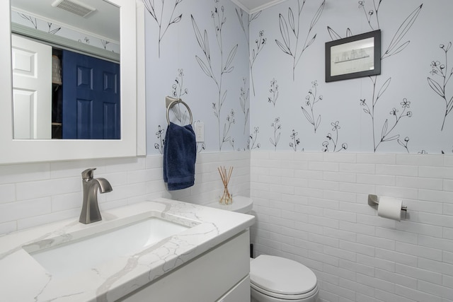 half bath with toilet, a wainscoted wall, vanity, visible vents, and tile walls