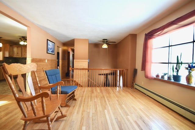 living area with light wood finished floors, a baseboard radiator, a ceiling fan, wainscoting, and an upstairs landing