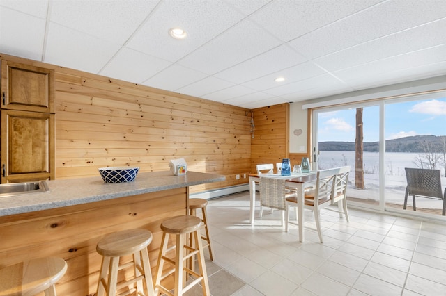 dining space featuring wood walls, baseboard heating, a drop ceiling, and light tile patterned flooring