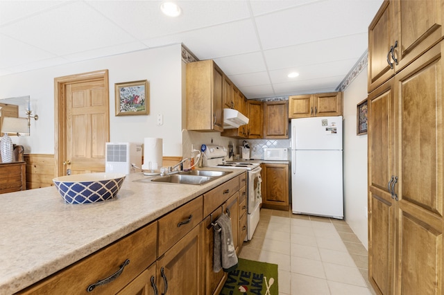 kitchen featuring under cabinet range hood, white appliances, a sink, light countertops, and brown cabinets