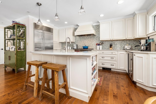 kitchen with premium range hood, white cabinetry, backsplash, stainless steel built in fridge, and an island with sink