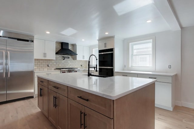 kitchen featuring high end fridge, white cabinets, a sink, wall chimney range hood, and an island with sink