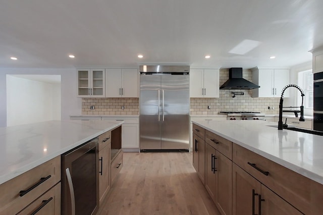 kitchen featuring stainless steel appliances, glass insert cabinets, white cabinetry, a sink, and wall chimney exhaust hood