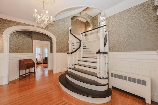 stairway with wood finished floors, wainscoting, radiator heating unit, and wallpapered walls