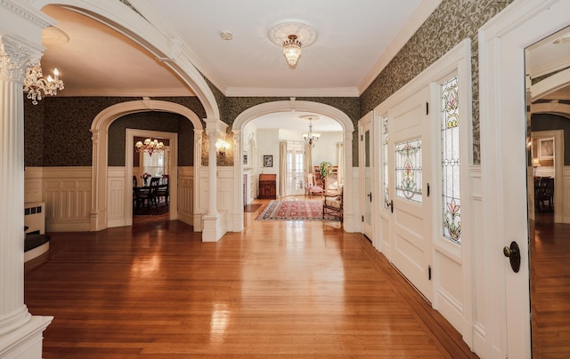 entrance foyer featuring wallpapered walls, arched walkways, wainscoting, an inviting chandelier, and ornate columns