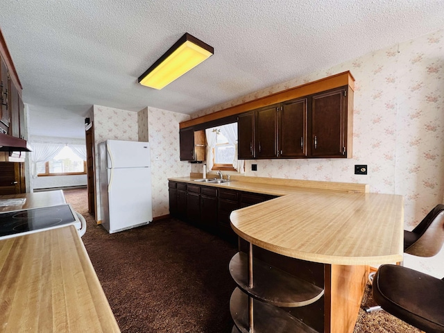 kitchen with wallpapered walls, freestanding refrigerator, a peninsula, dark colored carpet, and a textured ceiling