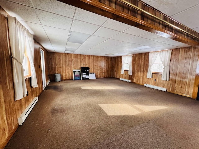 carpeted empty room with a baseboard radiator, wooden walls, and a drop ceiling